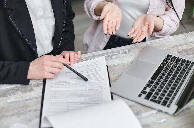 a person holding black pen pointing something on a paper discussing with someone else in fro
nt of a computer