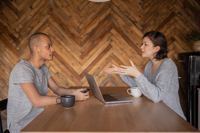 two-people-sitting-at-a-wooden-table-having-a-conversation-with-a-laptop-and-a-mug-in-front-of-them